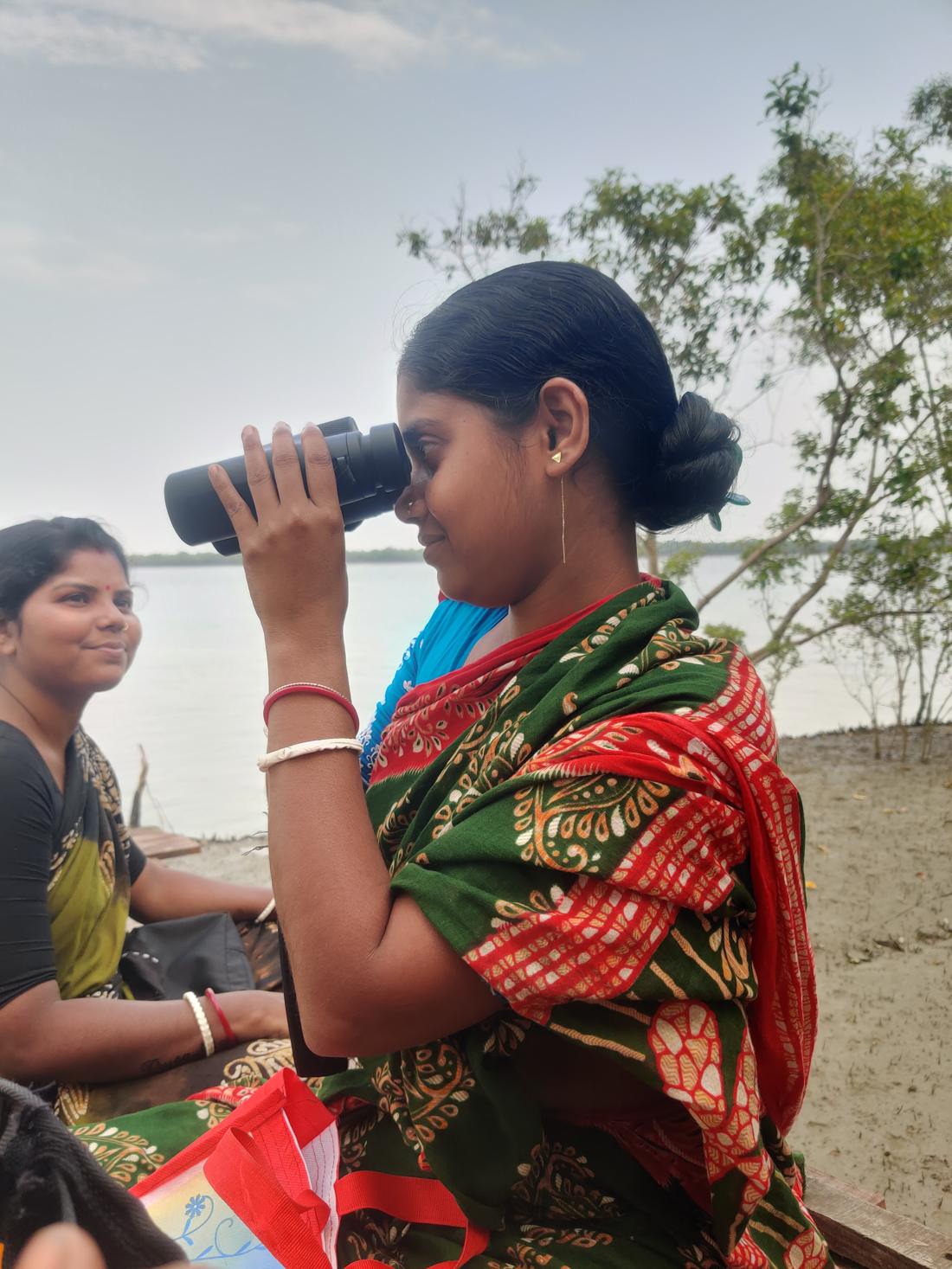 A young woman looks through binoculars on the riverbank while another smiles beside her.