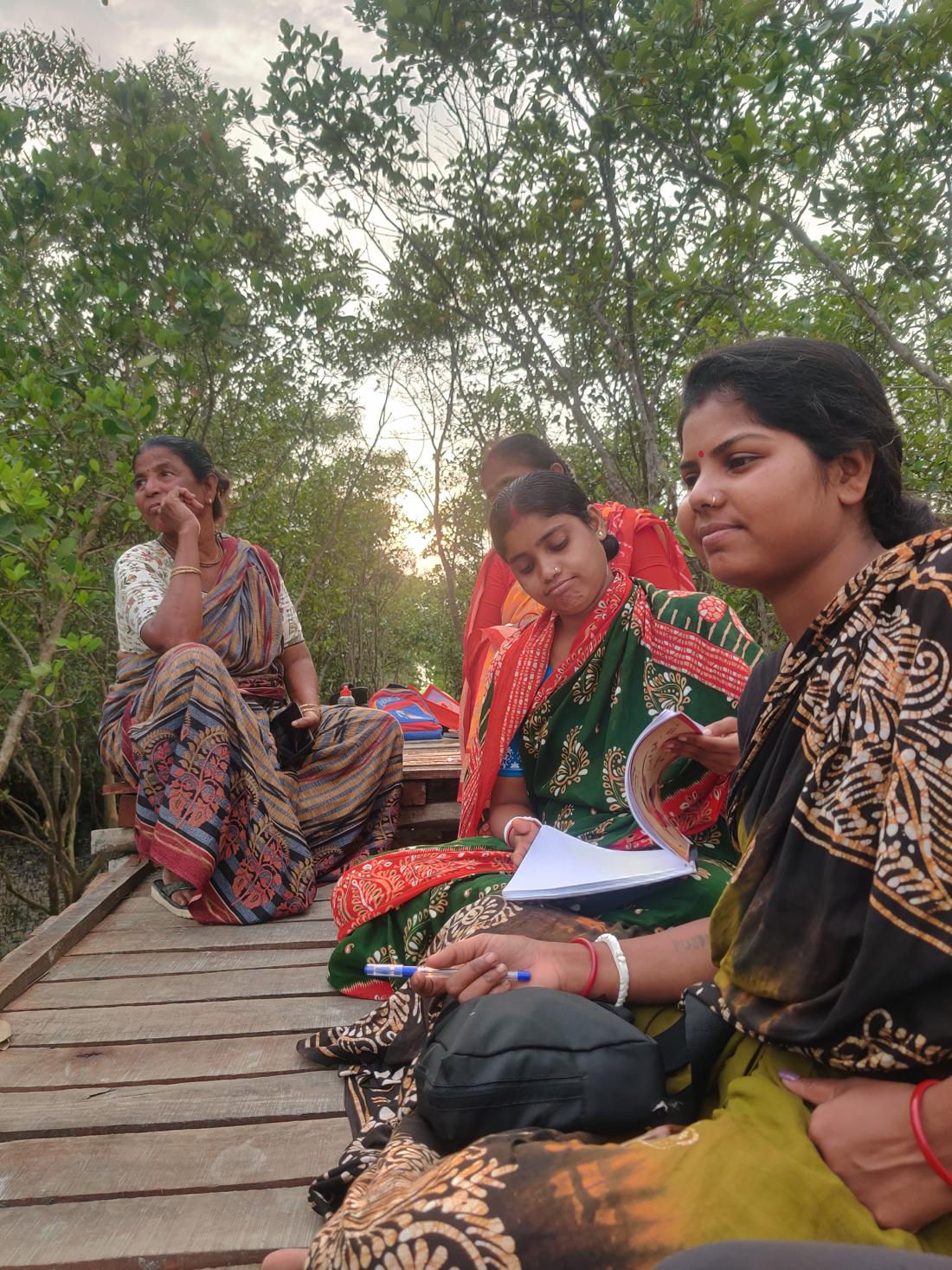 Women sitting on a wooden jetty among mangroves with notebooks and a field guide.