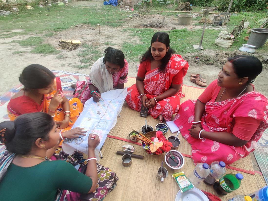 Five women seated on a mat outdoors, painting and drawing together.